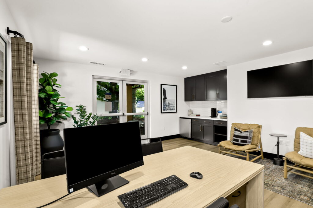 Interior entryway with double glass doors, potted plants, desk with computer, and a kitchenette with dark cabinets and a small refrigerator.