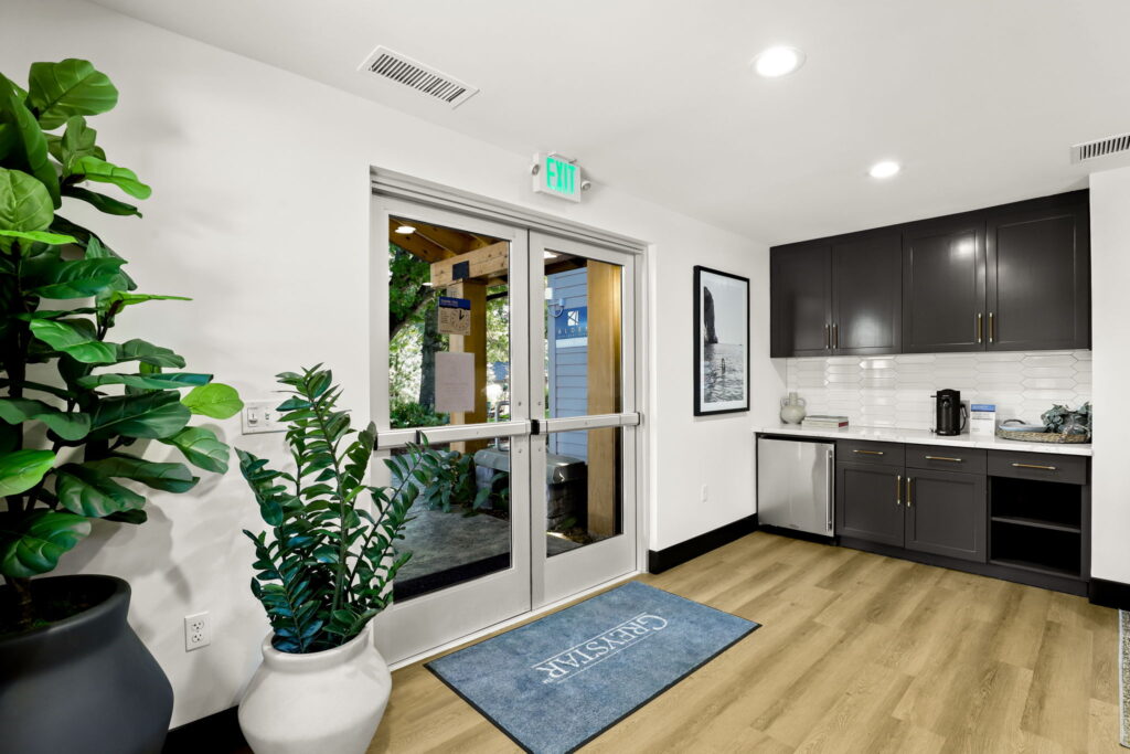 Interior entryway with double glass doors, potted plants, a blue rug, and a kitchenette with dark cabinets and a small refrigerator.